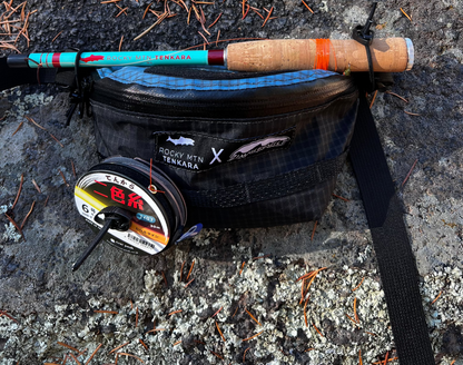 A Tenkara fishing rod with a turquoise and red handle, lying next to a black hip pack labeled 'ZimmerBuilt x Rocky MTN Tenkara' and various fishing accessories.